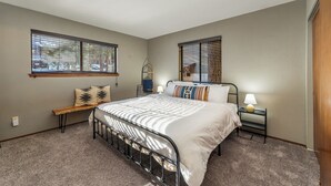 Un dormitorio con cama, dos ventanas, un banco, una mesita de noche y una lámpara.