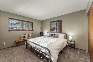 A bedroom with a bed, two windows, a bench, a nightstand, and a lamp.