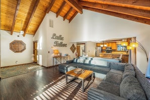 A spacious living room with a large gray sectional sofa, a wooden coffee table, and a stone-patterned rug. A kitchen is visible in the background with wooden cabinets and a hanging light fixture.