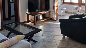 Un soggiorno con una TV a schermo piatto su un supporto in legno, un tavolino da caffè con piano in vetro, un divano scuro e una poltrona in vimini.