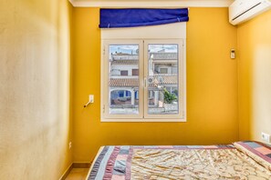 A room with yellow walls, a bed with a patterned bedspread, a window with a view of buildings, and an air conditioning unit. - Holiday Home 'Apartmento Girelle' with Sea View, Wi-Fi and Air Conditioning (L'Escala)