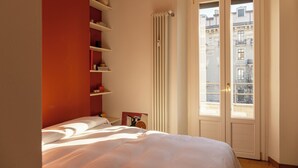 Un dormitorio con cama, radiador, ventana con vista a edificios y estantes con objetos.