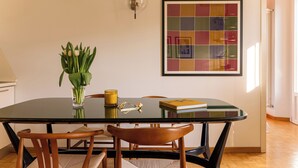 Un comedor con una mesa de vidrio, sillas de madera, un jarrón con hojas verdes y una obra de arte enmarcada en la pared.