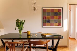 Un coin repas avec une table en verre, des chaises en bois, un vase contenant des feuilles vertes et une œuvre d’art encadrée accrochée au mur.