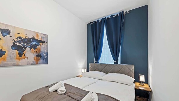 Una cama bien hecha con un mapa mundial en la pared, una cortina azul oscuro y una mesita de noche con una lámpara.