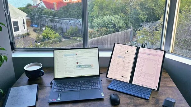 Ein Arbeitsplatz mit zwei Laptops, einer Maus und einer Tasse auf einem Schreibtisch an einem Fenster mit Blick auf einen See und Hügel.