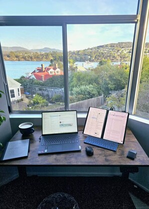 Een werkplek met twee laptops, een muis en een beker op een bureau, met uitzicht op een meer en heuvels.