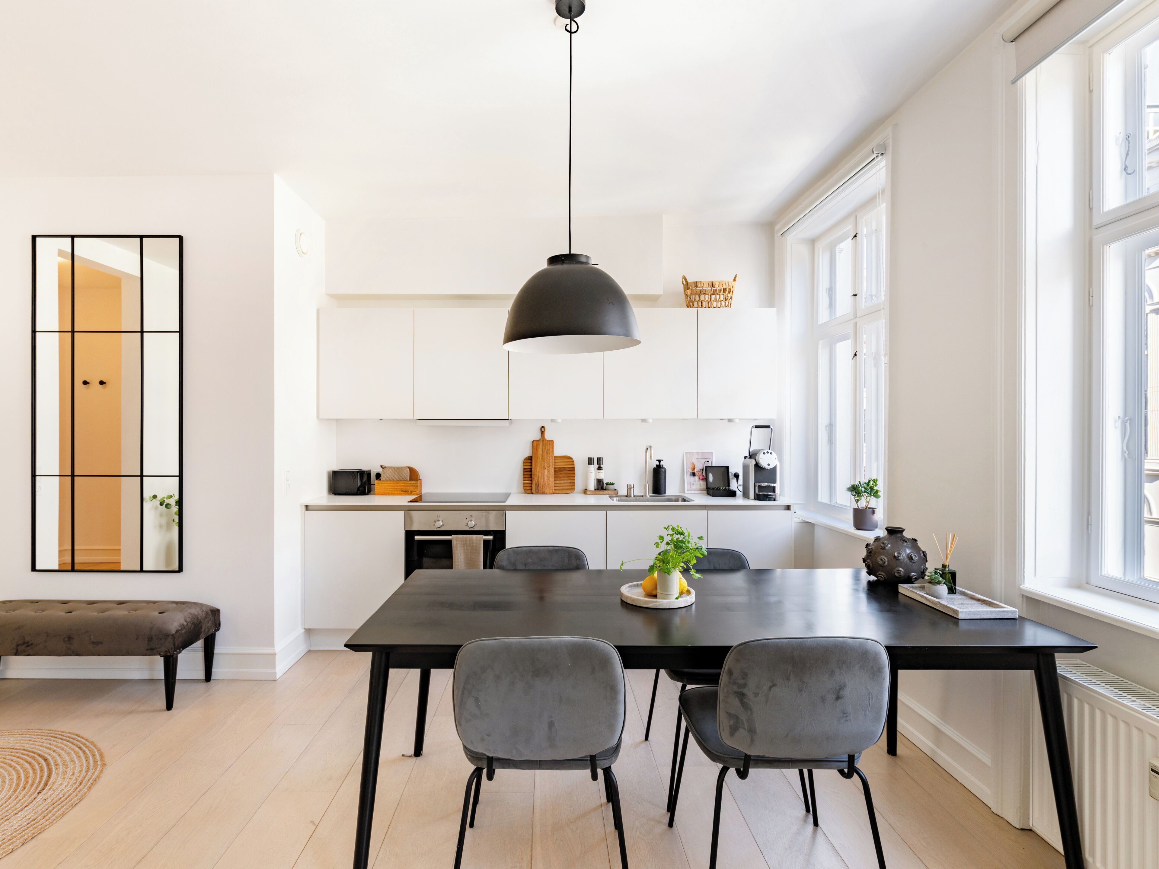 A modern kitchen with a dining area, featuring a large table, chairs, a hanging light fixture, and a window with natural light.