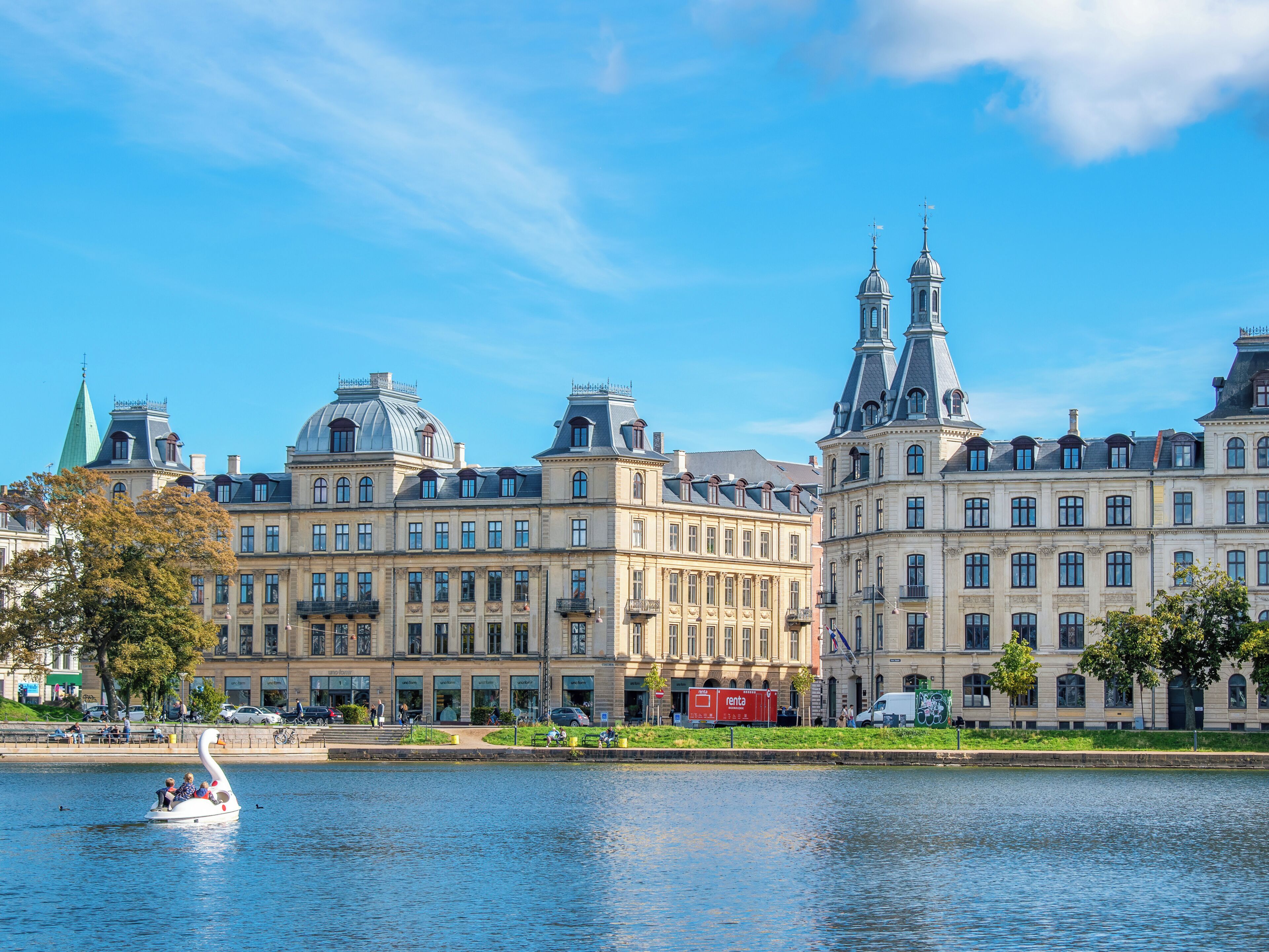 A historic building with classical architecture, a river in the foreground, and a swan-shaped boat with people.