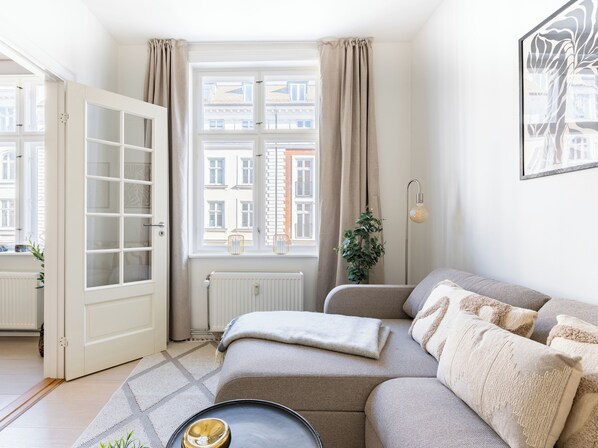 A living room with a sofa, a radiator, a window with curtains, a door, and a plant. - Shield 2 - Great Apartment in the Centre of Copenhagen (Copenhagen)