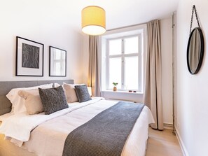 A neatly arranged bedroom with a bed, two framed artworks, a window with curtains, and a hanging mirror. - Shield 2 - Great Apartment in the Centre of Copenhagen (Copenhagen)