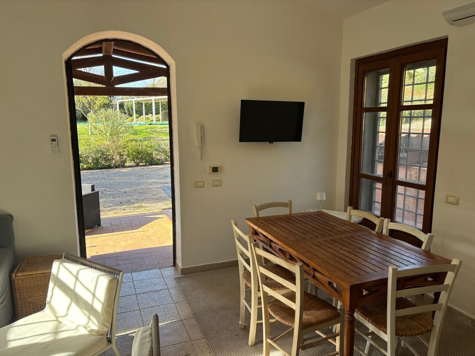 A dining area with a wooden table and chairs, a television mounted on the wall, and an arched doorway leading to an outdoor area.