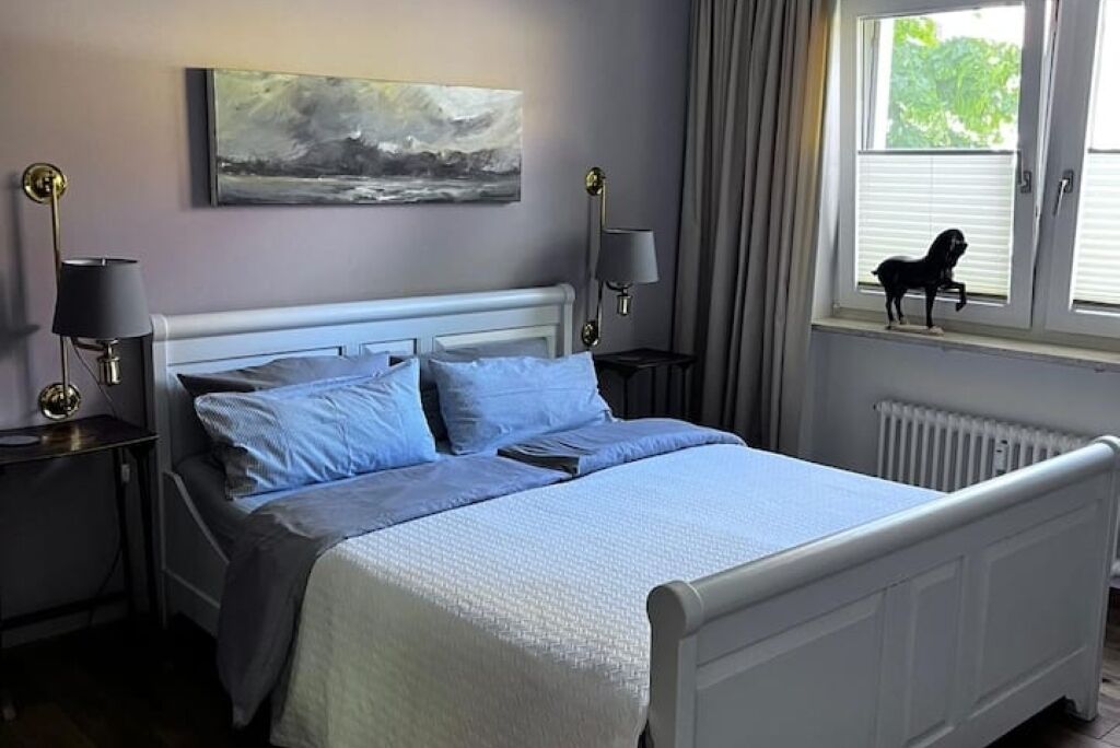 A neatly made bed with a white headboard, two bedside lamps, a painting on the wall, and a window with a view of greenery.