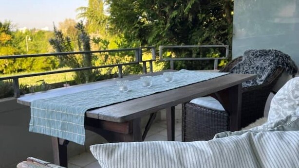 Un balcón con una mesa para dos, una silla de mimbre y vistas al verdor.