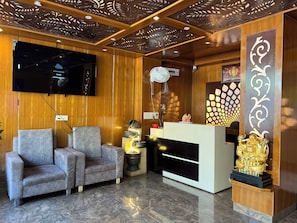 A modern reception area with a wooden ceiling, a flat-screen TV, two grey armchairs, a reception desk, and a decorative golden statue. - The White Rain Hotel (Lansdowne)