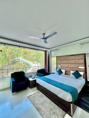 A modern hotel room with a large bed, a ceiling fan, a seating area with a chair, and a view of trees and a bridge outside. - The White Rain Hotel (Lansdowne)