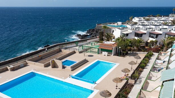 Tagpoolområde med to swimmingpools, omgivet af liggestole og parasoller, med udsigt over havet.