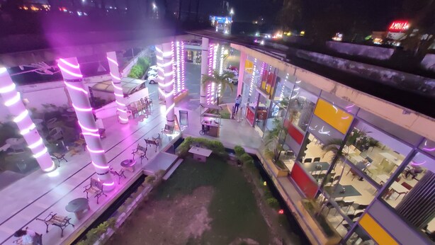 An outdoor area with seating, a small garden, and a building with large windows and a yellow section. - EXPRESS FOOD PLAZA (Kolaghat)