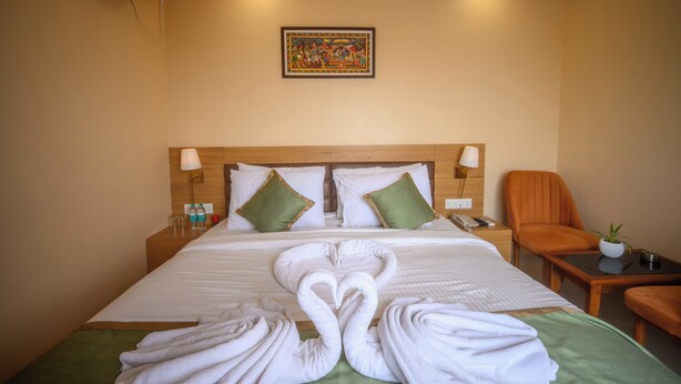 A hotel room with a large bed, two pillows, a nightstand, a chair, a small table, a wall-mounted air conditioner, and a painting on the wall.