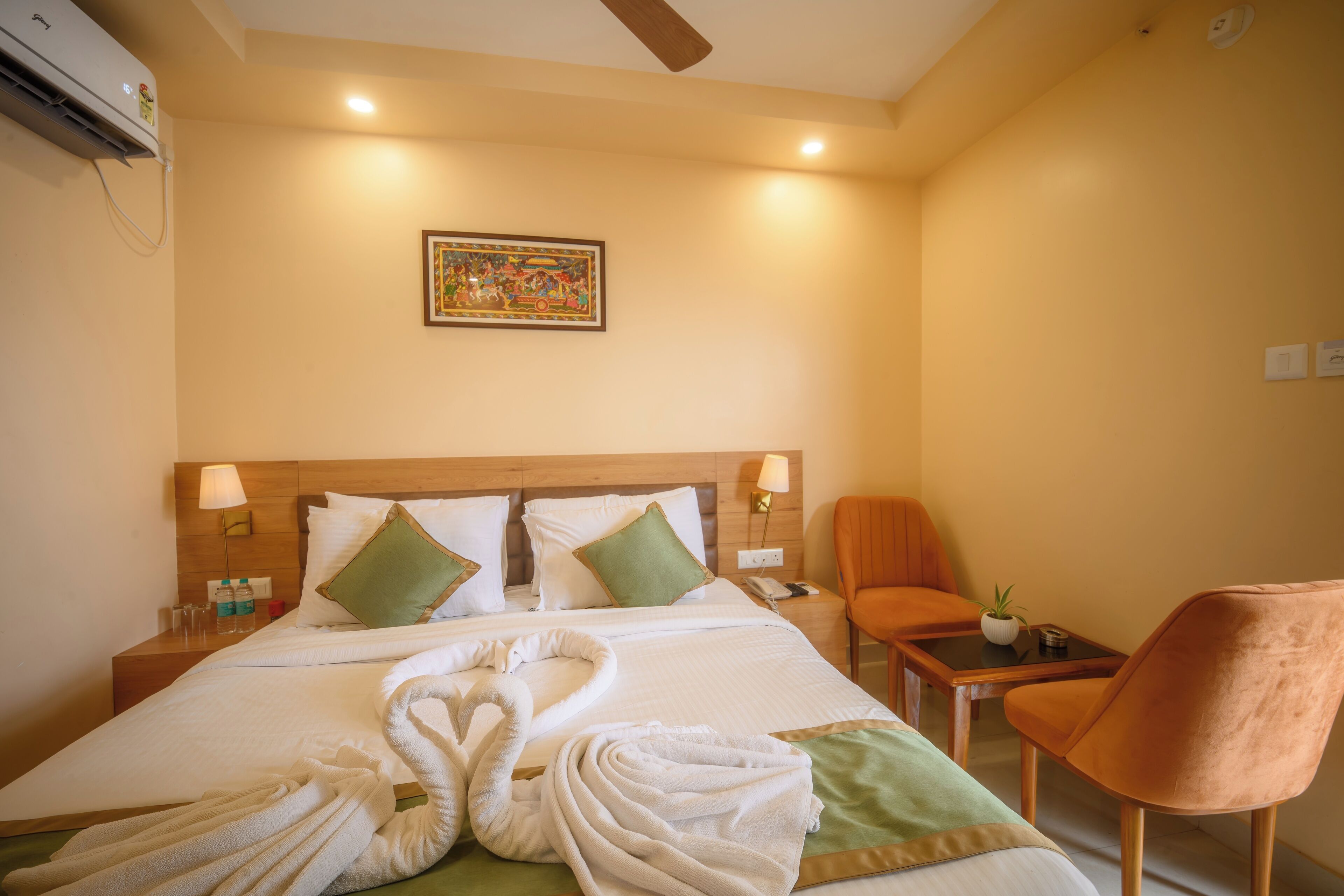Habitación de hotel con cama, dos almohadas, mesita de noche, lámpara, silla, pequeña mesa, aire acondicionado de pared y un cuadro en la pared.