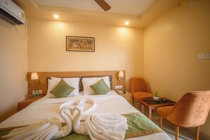 A hotel room with a bed, two pillows, a nightstand, a lamp, a chair, a small table, a wall-mounted air conditioner, and a framed picture on the wall. - WORMWOOD SYNERGY LA BELLA (Brahmagiri)