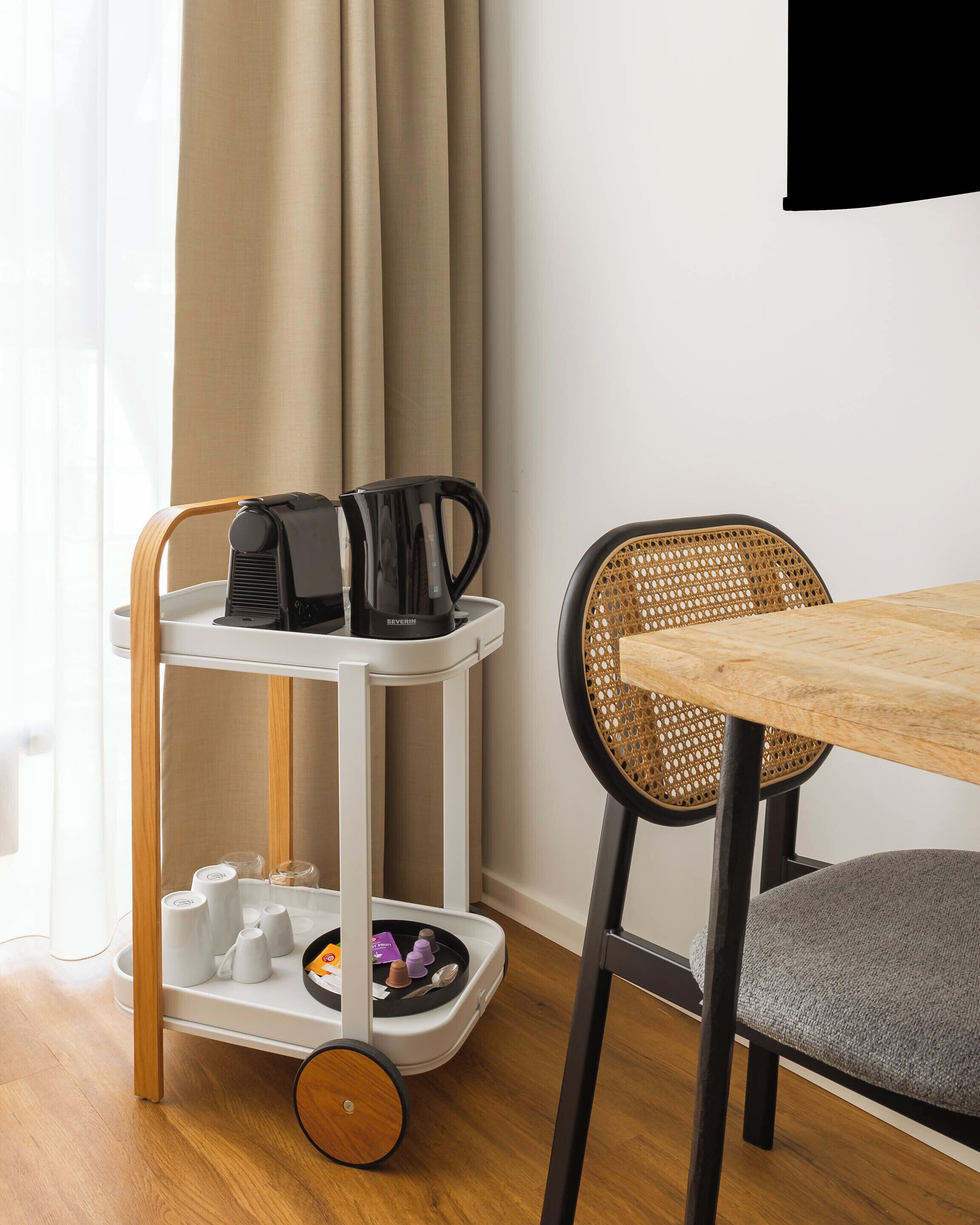 A room with a wooden table, a wicker chair, a white cart with a coffee maker, and a curtain.