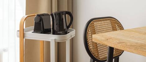 A room with a wooden table, a wicker chair, a white cart with a coffee maker, and a curtain.