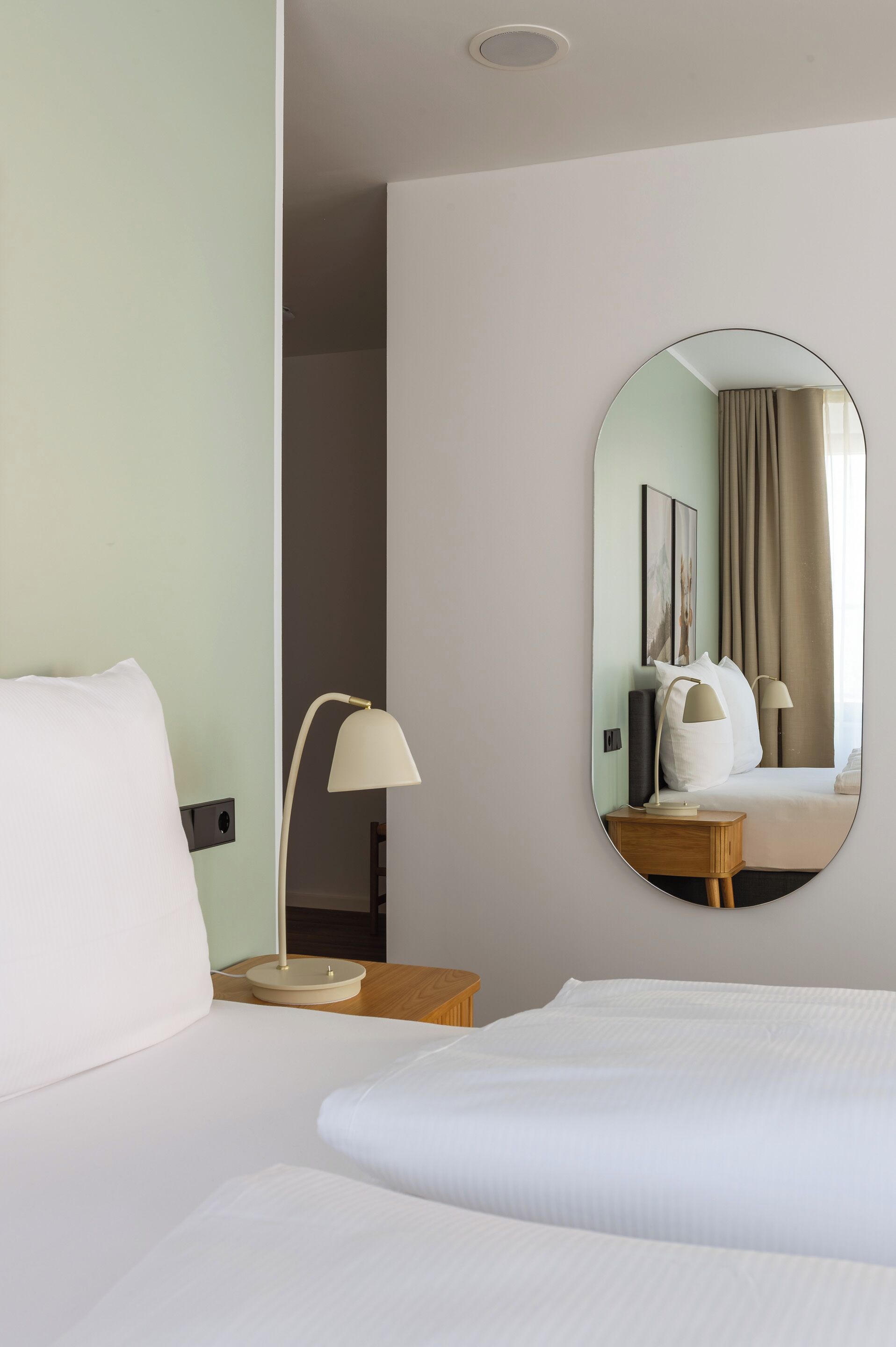 A hotel room with a bed, a bedside lamp, a nightstand, a mirror, and a door leading to another room.