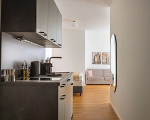 A modern kitchen with a stove, coffee maker, and cabinets, leading to a living area with a sofa and artwork on the wall. - Limehome Leverkusen Wiesdorfer Platz (Leverkusen)