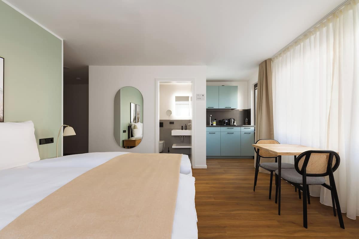A hotel room with a bed, a desk, a chair, a mirror, a kitchenette, and a bathroom.