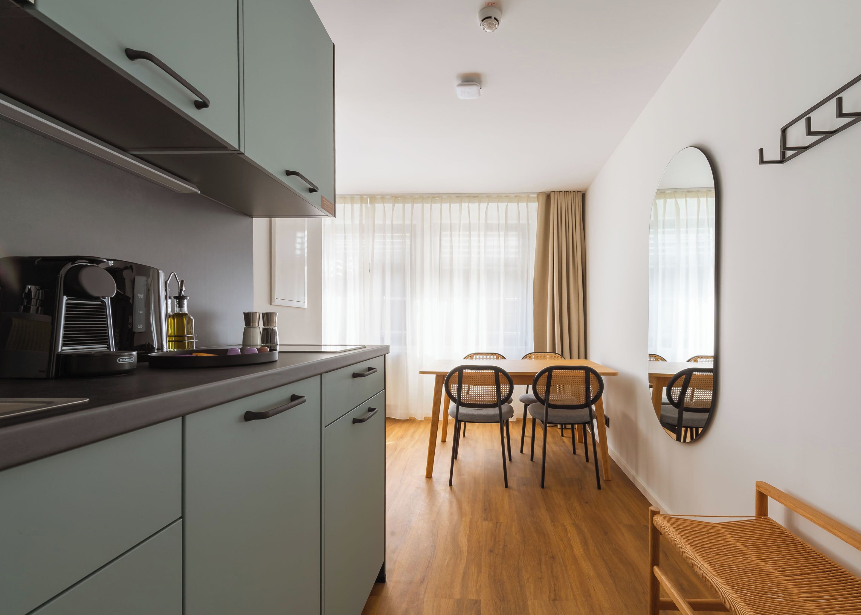 A modern kitchen with a coffee machine, a dining table with chairs, and a large mirror on the wall.
