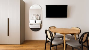 Una habitación moderna con una mesa redonda de madera, cuatro sillas grises tapizadas, un sofá blanco, un espejo grande y un televisor de pantalla plana montado en la pared.