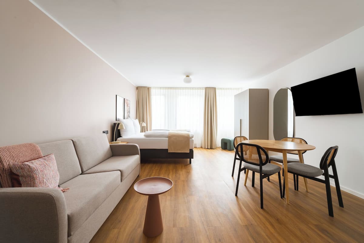 A modern hotel room with a sofa, a dining table with chairs, a TV mounted on the wall, and a large window with curtains.