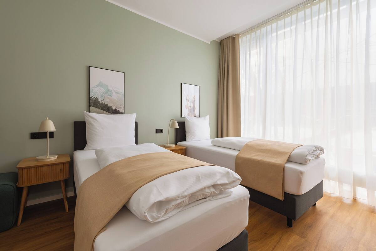 A hotel room with two beds, a nightstand with a lamp, a wall-mounted picture, and a large window with sheer curtains.