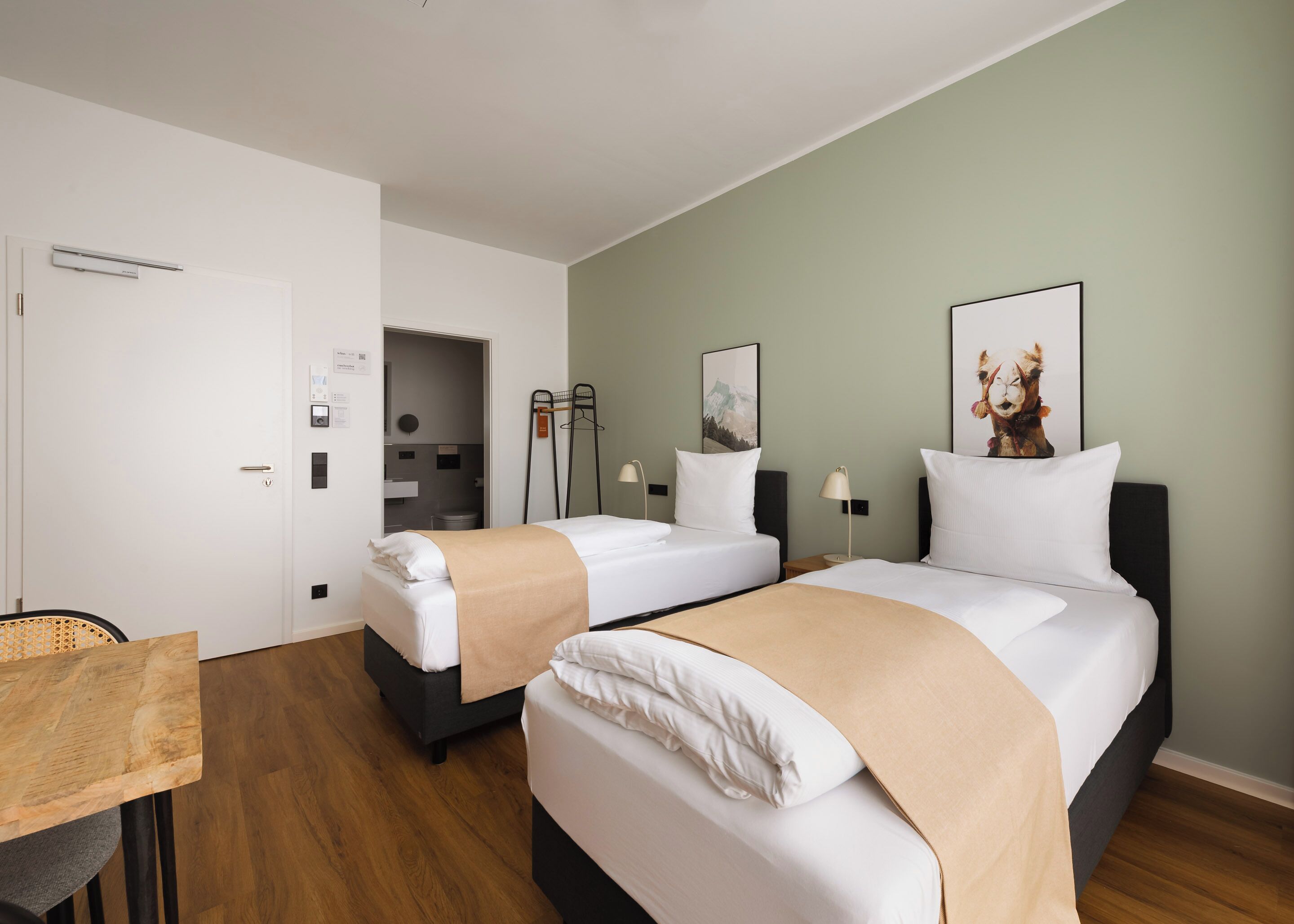 Quarto de hotel com duas camas, uma mesa de madeira, uma cadeira, escrivaninha, abajur e um quadro na parede.