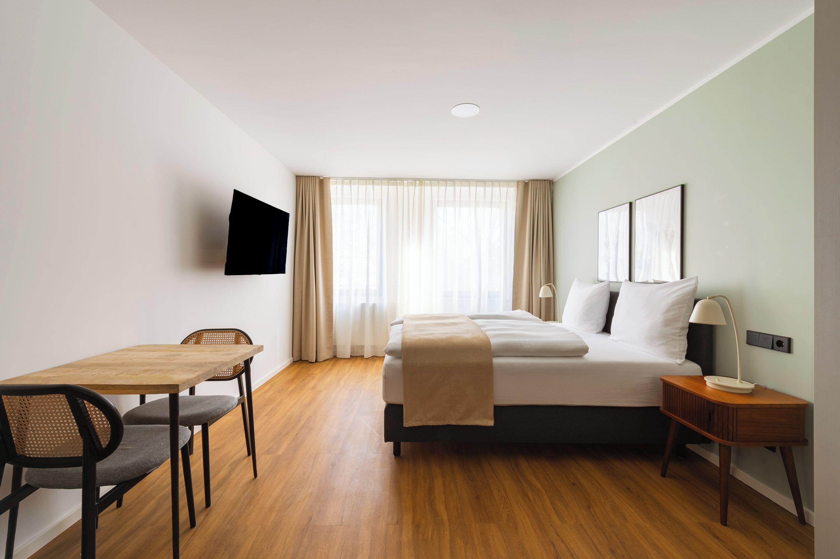 Una camera d'albergo con un letto, una scrivania, una sedia, una TV a muro e una finestra con tende.