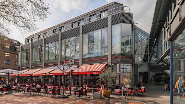 Un café al aire libre con mesas y sillas, ventanas amplias y una fachada de edificio moderna.