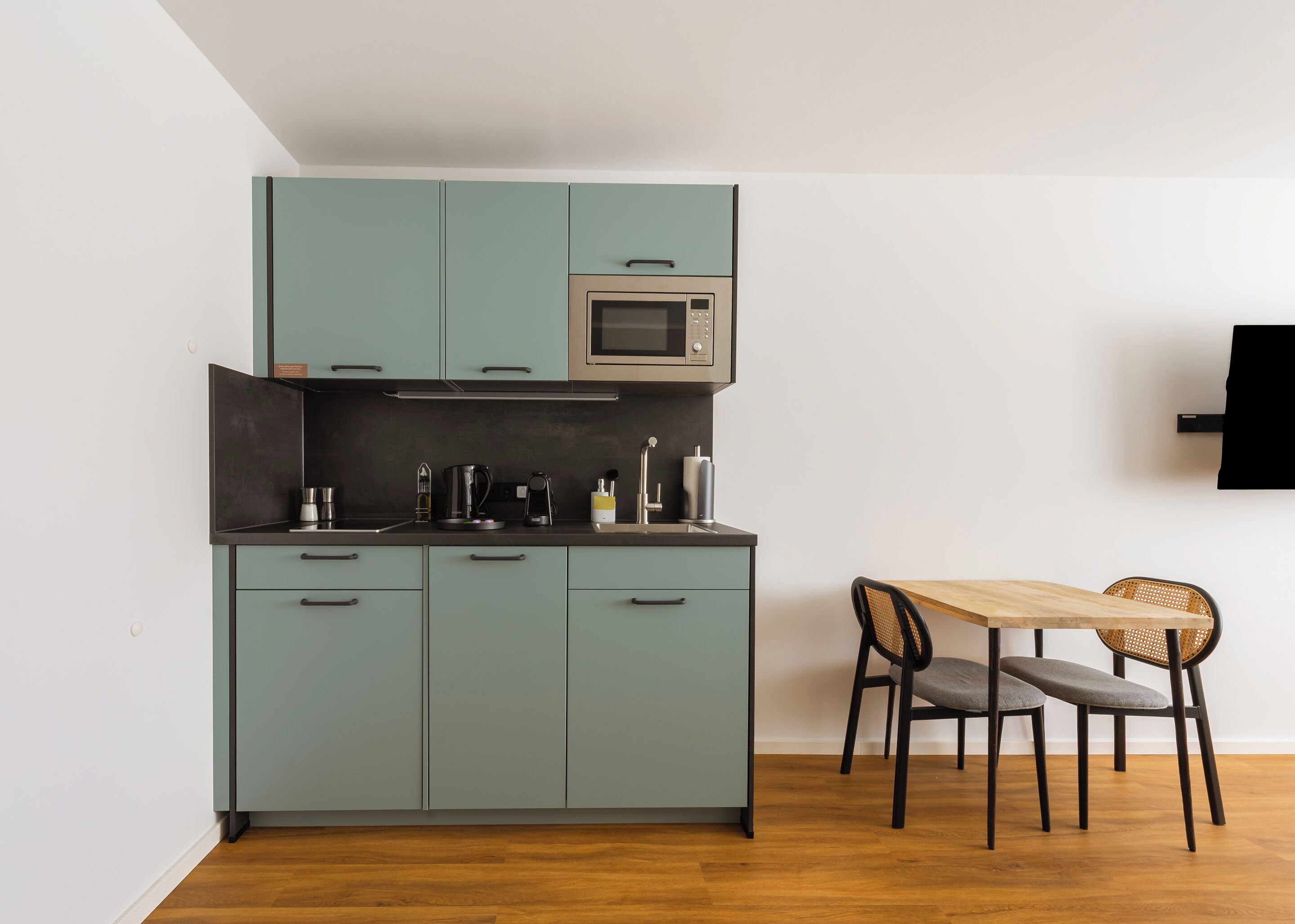 A compact kitchen with teal cabinets, a microwave, and a dining area with a wooden table and two chairs.