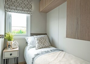 A bedroom with a bed, bedside table, window with curtains, and a view of greenery outside.