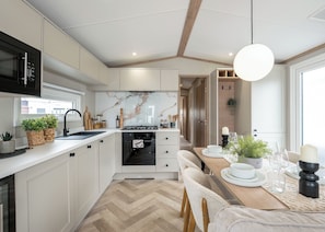 A modern kitchen with white cabinets, a herringbone floor, a built-in oven, and a dining area with a wooden table and chairs. - Premium Hot Tub 4 bedroom Holiday Home sleeps 8 (Hayle)
