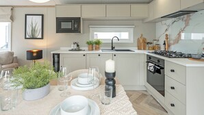 Une cuisine moderne équipée d’un four à micro-ondes, d’une cuisinière et d’un four encastrés. On y trouve un coin repas avec une table dressée pour deux personnes, une plante en pot et une bougie.