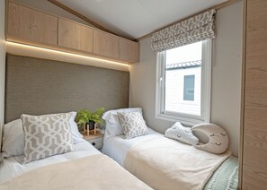 A compact bedroom with two beds, a window with a view, and built-in wooden cabinets. - Premium Hot Tub 4 bedroom Holiday Home sleeps 8 (Hayle)