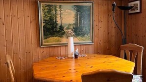 Un coin repas comprenant une table en bois, des chaises, un tableau encadré représentant une forêt et un lampadaire.