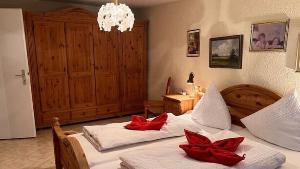 Une chambre d’hôtel avec une armoire en bois, un lit avec du linge de maison blanc et des serviettes rouges, une table de chevet avec une lampe, et des tableaux accrochés au mur.