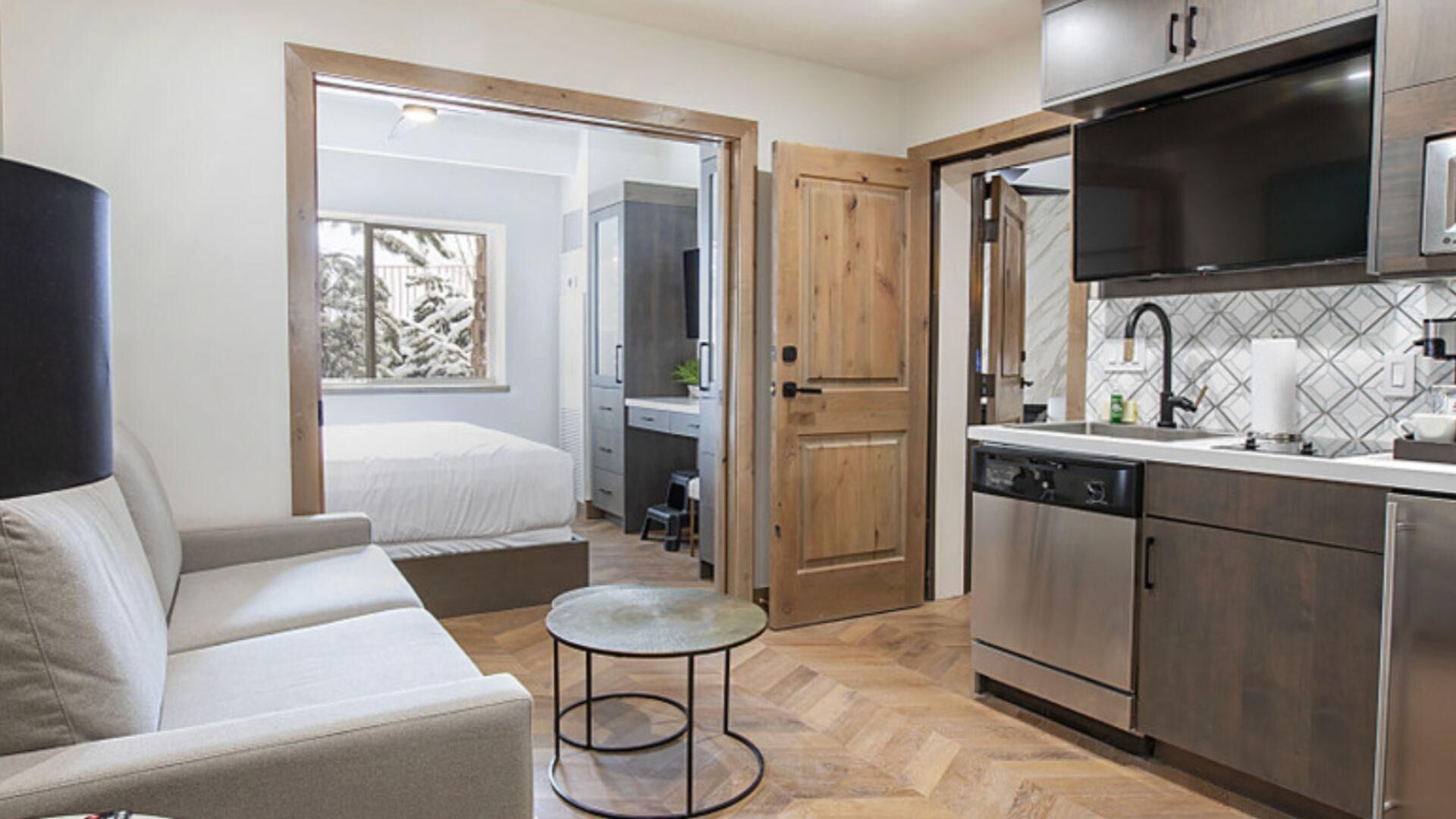 Un espacio habitable compacto con sofá, una mesita redonda, una kitchenette equipada con electrodomésticos modernos y una puerta que da a un dormitorio con vistas a árboles cubiertos de nieve.