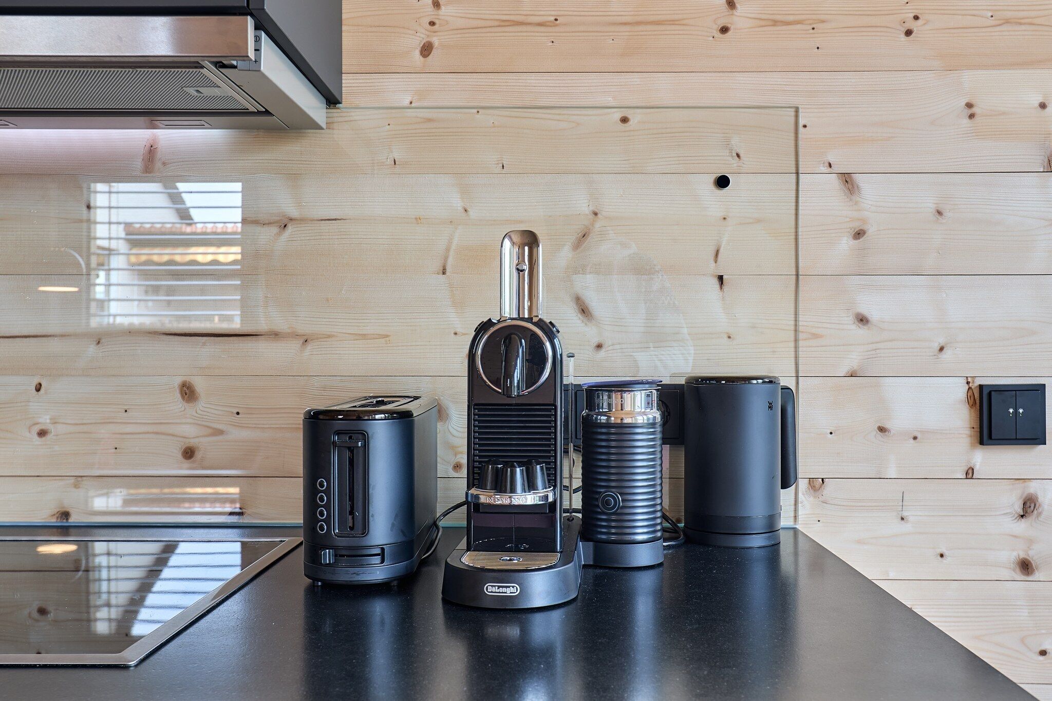 Una cocina con diseño moderno, que cuenta con una campana extractora de acero inoxidable, una cafetera, un tostador y un hervidor sobre una encimera negra frente a una pared de madera.