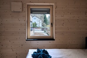 Huone, jossa on puinen seinä, valkoisella lakanalla päällystetty sänky ja ikkuna, josta avautuu näkymä kadulle ja rakennuksiin.