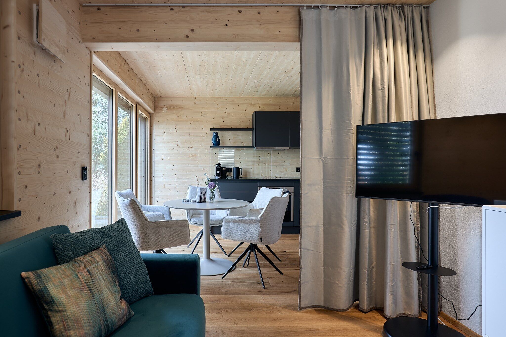 A modern living room with a sofa, chairs, a TV, and a kitchen in the background.