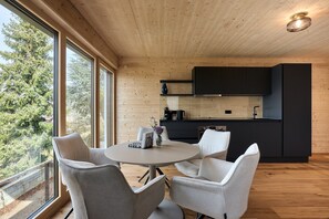 Moderni keittiö, jossa on tummat kaapit, pyöreä pöytä tuoleineen ja suuret ikkunat, joista avautuu näkymä puita kohti.