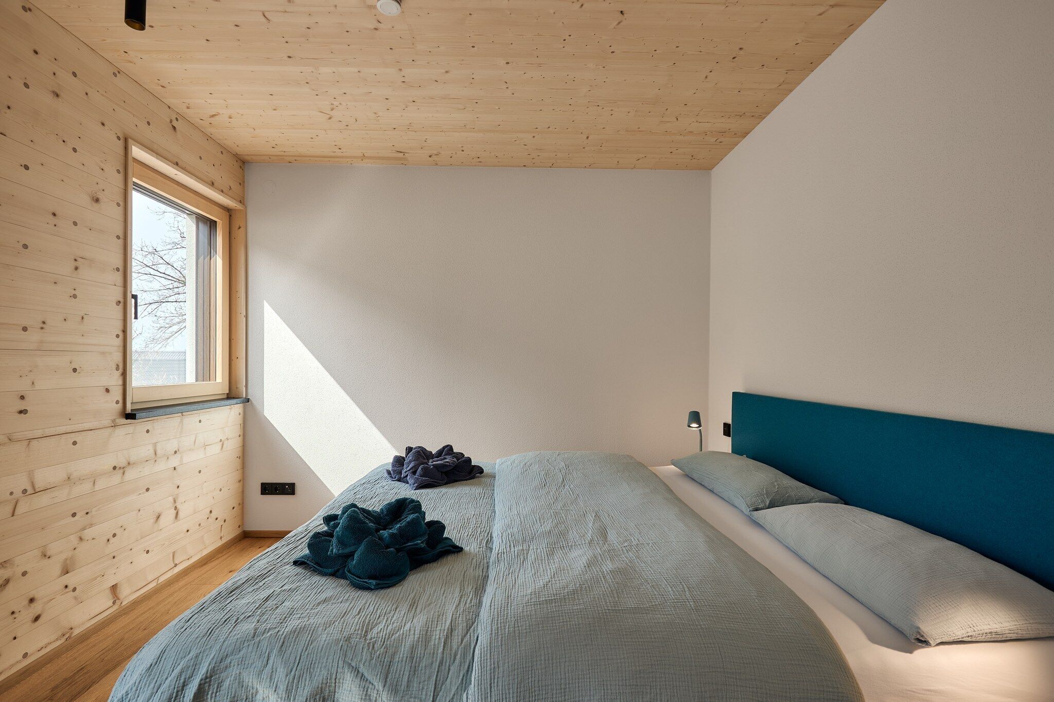 Ein Schlafzimmer mit schrägen Wänden, Holztäfelung, einem Bett mit blauer Kopfteil und einem Fenster, das natürliches Licht hereinlässt.